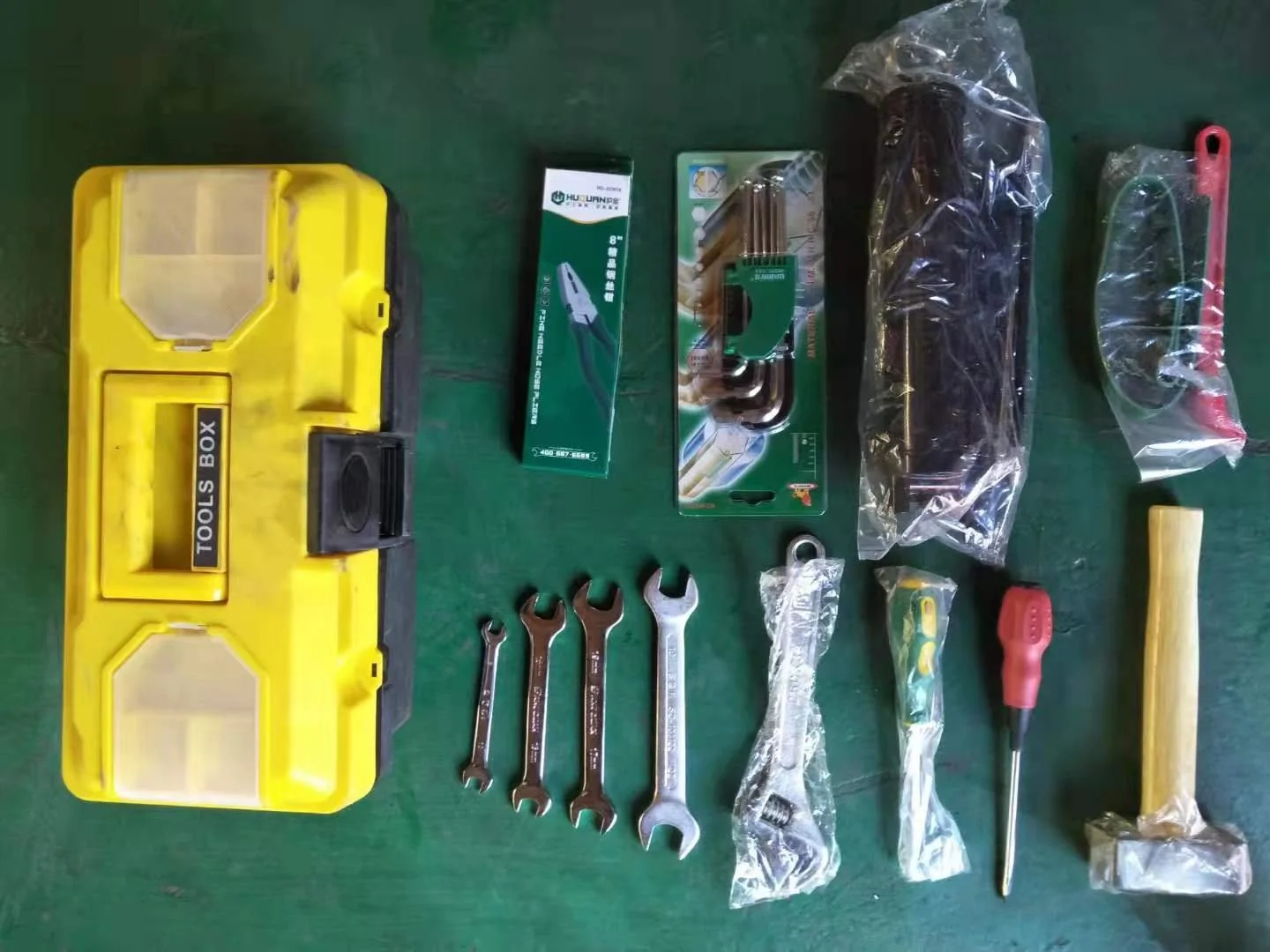 A collection of tools and hardware laid out on a green surface, including a yellow toolbox, wrenches, screwdrivers, a spirit level, a box cutter, pliers, a hacksaw, a rubber mallet, and various hardware parts in plastic bags.